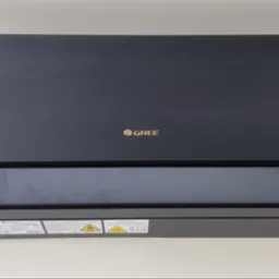 Czarna jednostka wewnętrzna klimatyzacji marki Gree, widoczna od frontu na jasnym tle, z naklejkami ostrzegawczymi w dolnej części obudowy.