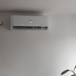 VENTUS - KLIMA Rafał Kućmierz - Biały klimatyzator ścienny marki Heiko z wyświetlaną temperaturą 26 stopni Celsjusza, zamontowany na białej ścianie w jasnym pomieszczeniu, obok lampki ściennej i zasłony w oknie.