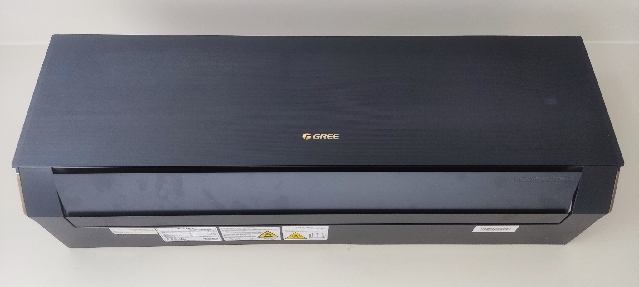 Czarna jednostka wewnętrzna klimatyzacji marki Gree, widoczna od frontu na jasnym tle, z naklejkami ostrzegawczymi w dolnej części obudowy.