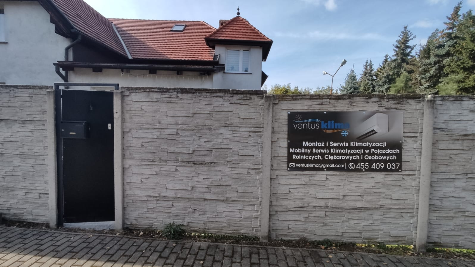 Tablica reklamowa firmy na ogrodzeniu z imitacji kamienia, oferującej montaż i serwis klimatyzacji, w tym mobilny serwis w pojazdach rolniczych, ciężarowych i osobowych.