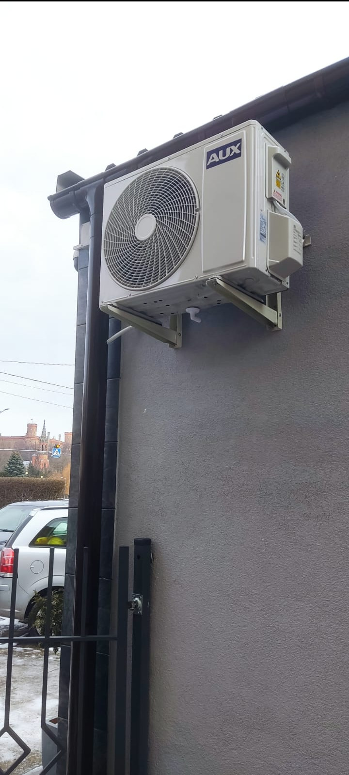 Zewnętrzna jednostka klimatyzacji marki AUX zamontowana na szarej ścianie budynku, z widocznym fragmentem dachu i rynny, w tle budynek z wieżami.