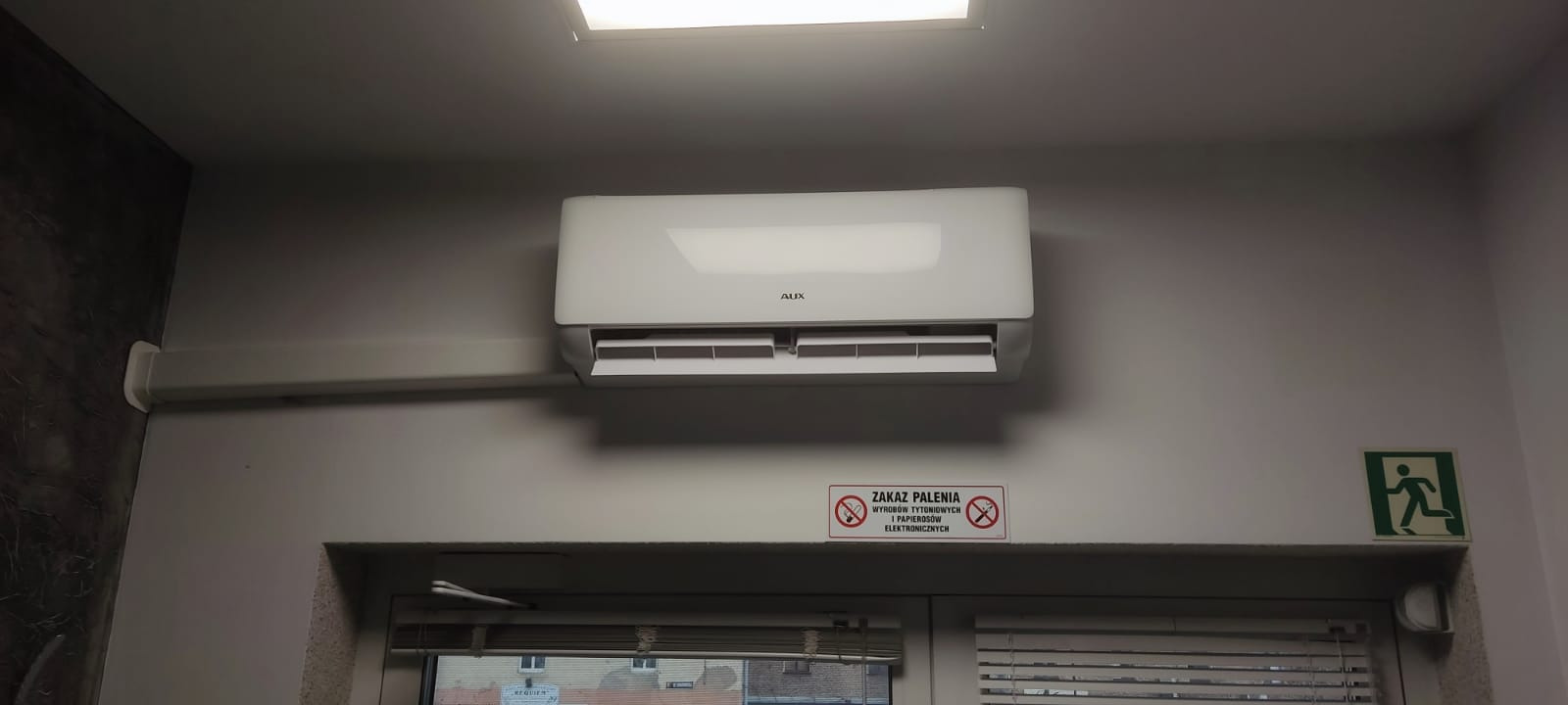 Biała klimatyzacja AUX zamontowana na ścianie nad oknem z żaluzjami, widoczny znak zakazu palenia i znak ewakuacyjny.