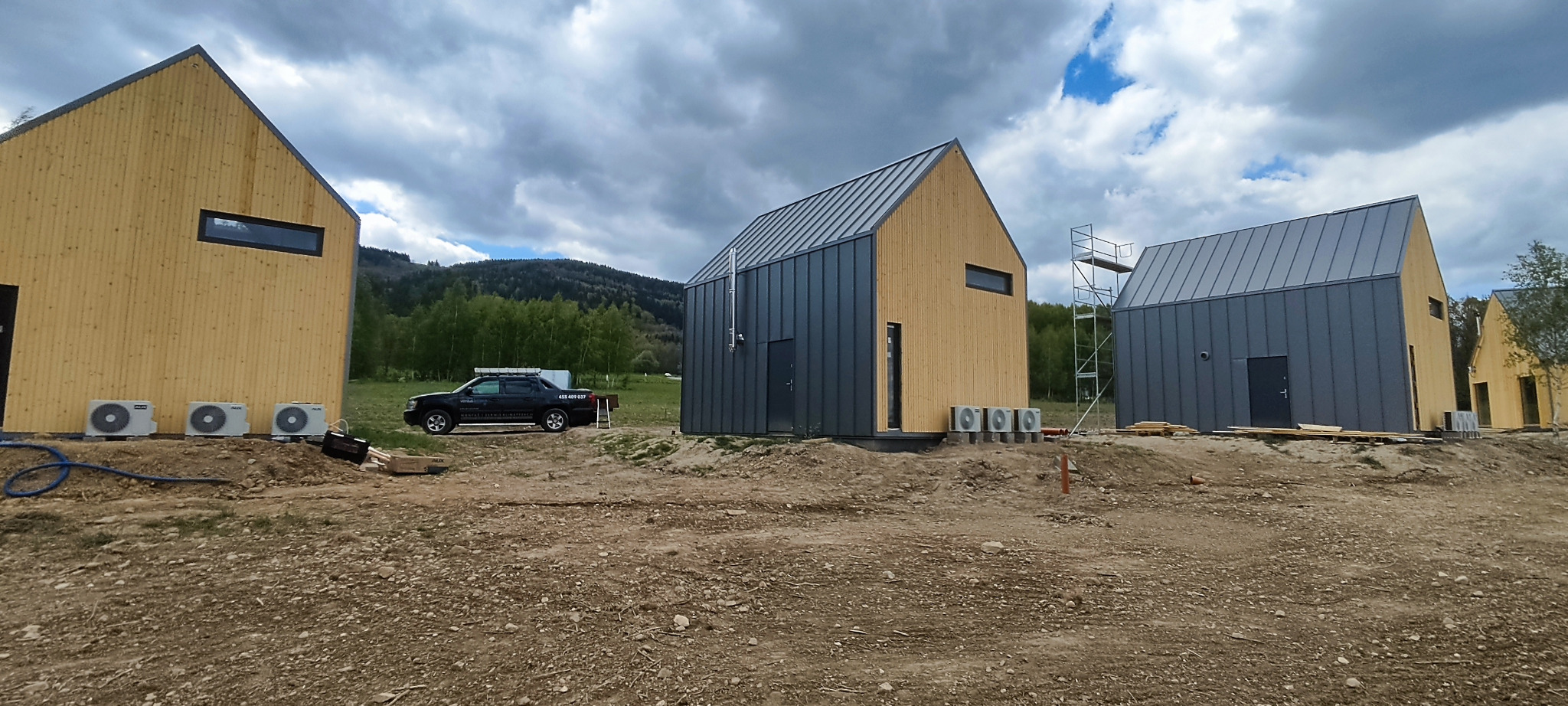 Nowoczesne domki modułowe z pompami ciepła AUX na zewnątrz, teren w trakcie zagospodarowania, w tle zalesione wzgórza i firmowy samochód.