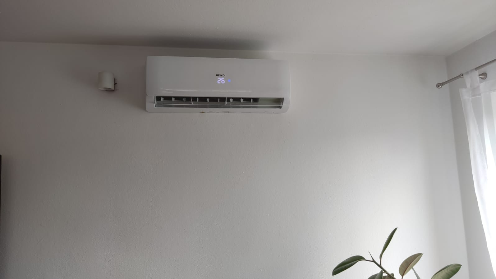 Biały klimatyzator ścienny marki Heiko z wyświetlaną temperaturą 26 stopni Celsjusza, zamontowany na białej ścianie w jasnym pomieszczeniu, obok lampki ściennej i zasłony w oknie.