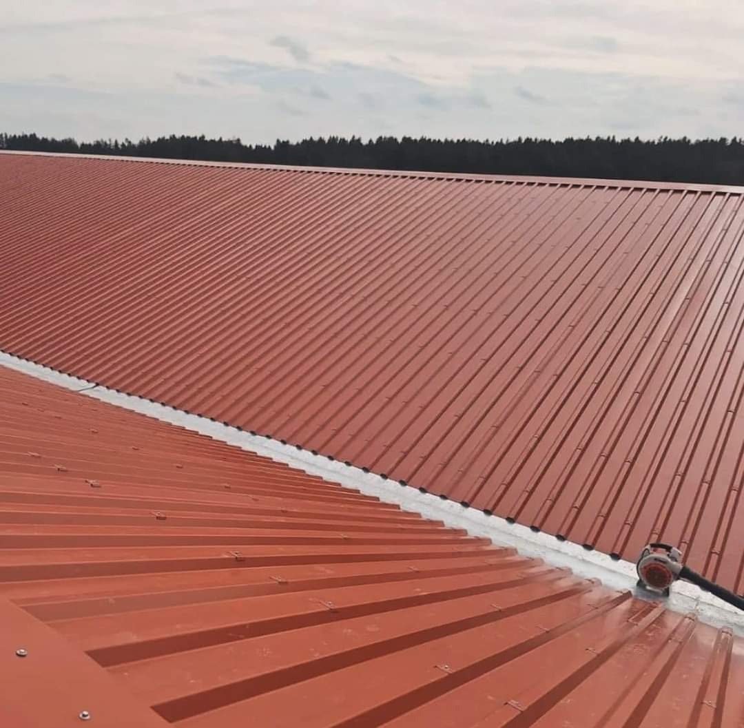 Czerwony dach z blachy trapezowej, widok z góry, z widocznym narzędziem do obróbki blachy przy krawędzi dachu i lasem w tle.