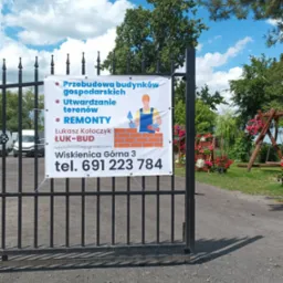 Ogłoszenie firmy budowlanej ŁUK-BUD zawieszone na metalowej bramie, oferującej przebudowę budynków gospodarskich, utwardzanie terenów oraz remonty, z ilustracją murarza i danymi kontaktowymi.