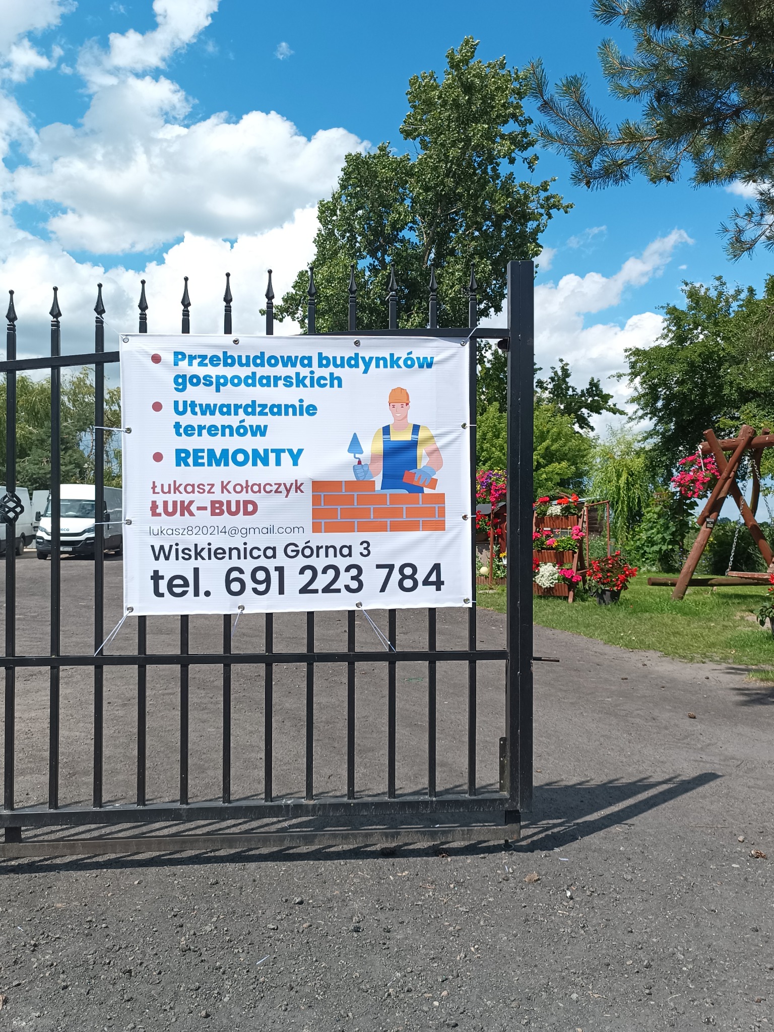 Ogłoszenie firmy budowlanej ŁUK-BUD zawieszone na metalowej bramie, oferującej przebudowę budynków gospodarskich, utwardzanie terenów oraz remonty, z ilustracją murarza i danymi kontaktowymi.