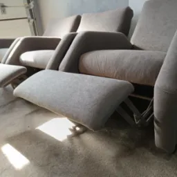 Rząd szarych, tapicerowanych foteli z wysuwanymi podnóżkami, ustawionych na betonowej podłodze, z widocznym mechanizmem rozkładania podnóżka.