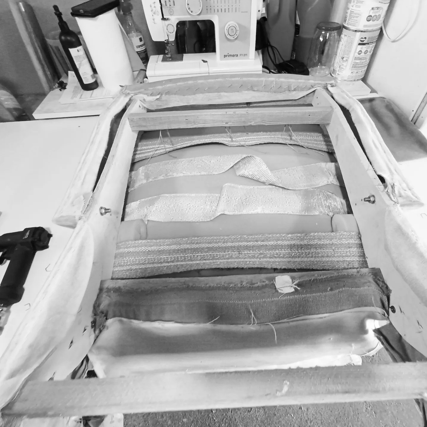 Fotel typu relax w trakcie renowacji: widoczny szkielet, pasy tapicerskie, zszywacz pneumatyczny i maszyna do szycia Primera w tle.