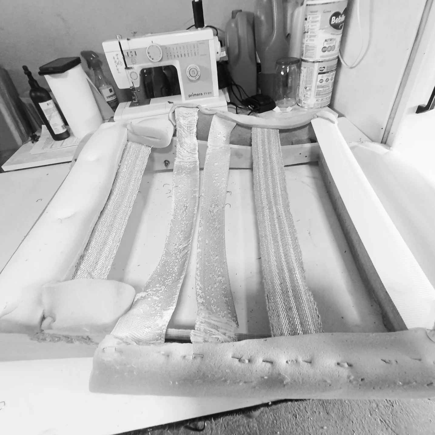 Czarno-białe zdjęcie ramy fotela w trakcie renowacji, z widocznymi pasami tapicerskimi, gąbką i zszywkami; w tle maszyna do szycia i inne akcesoria.
