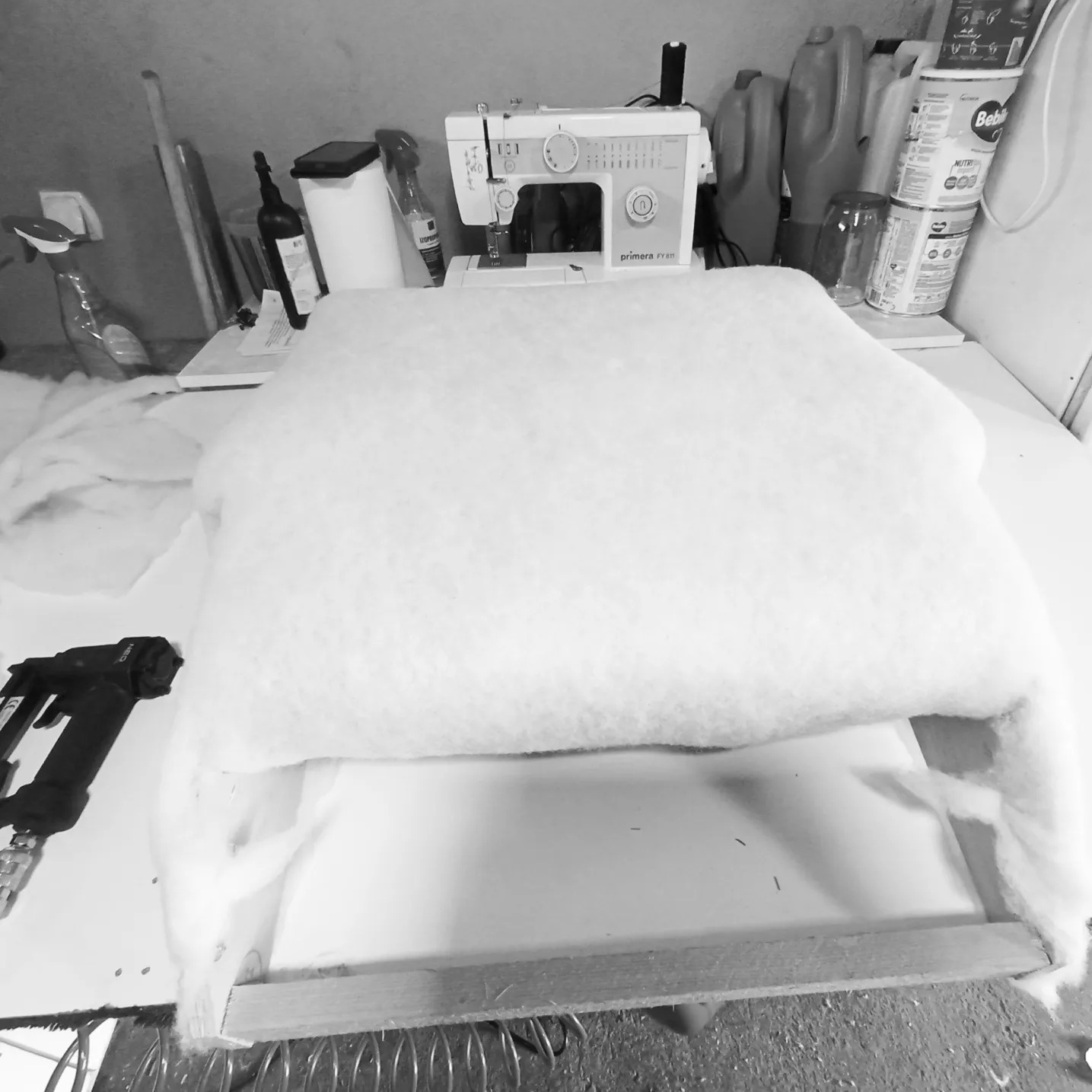 Fotel w trakcie renowacji: biała, puszysta warstwa waty tapicerskiej naciągnięta na drewniany szkielet siedziska, obok zszywacz tapicerski, w tle maszyna do szycia i akcesoria.