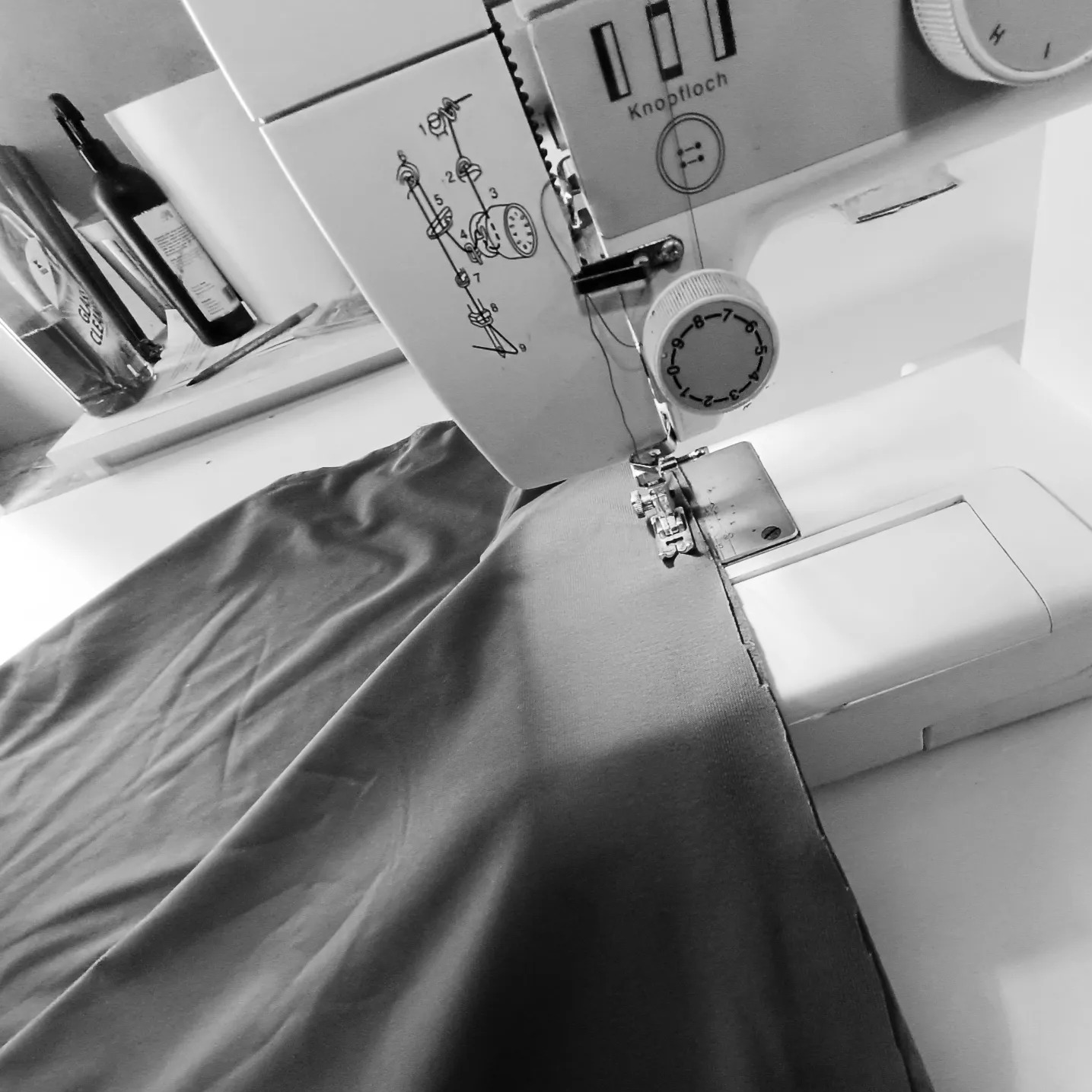 Czarno-białe zdjęcie maszyny do szycia w trakcie obróbki szwu na tkaninie, z widocznymi elementami maszyny, takimi jak pokrętło regulacji i mechanizm igły, oraz butelkami i długopisem w tle.