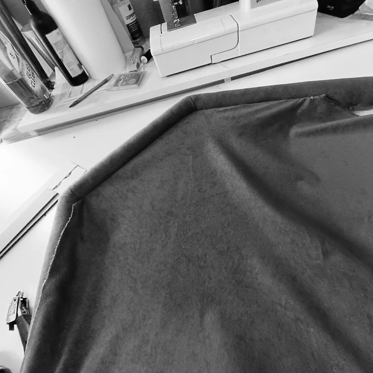 Fragment szarej tkaniny obiciowej z widocznym szwem, leżącej na stole obok maszyny do szycia i akcesoriów, w czarno-białej tonacji.