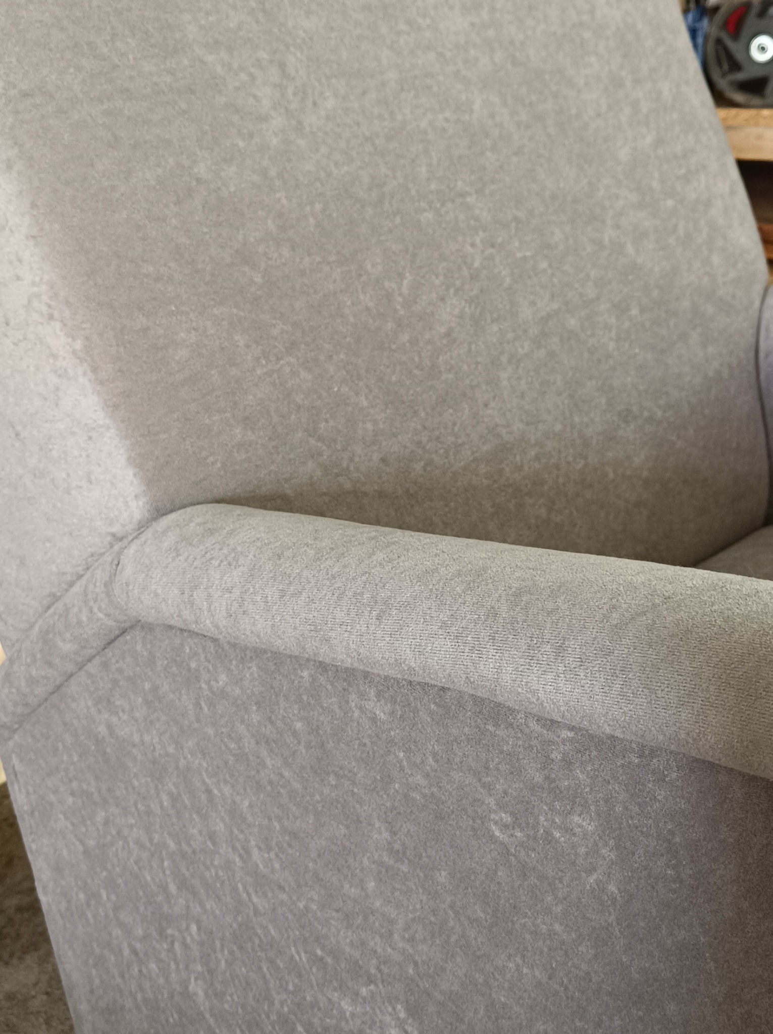 Fragment szarego fotela tapicerowanego z wyraźną fakturą materiału i zaokrąglonym podłokietnikiem, widoczne elementy drewniane w tle.