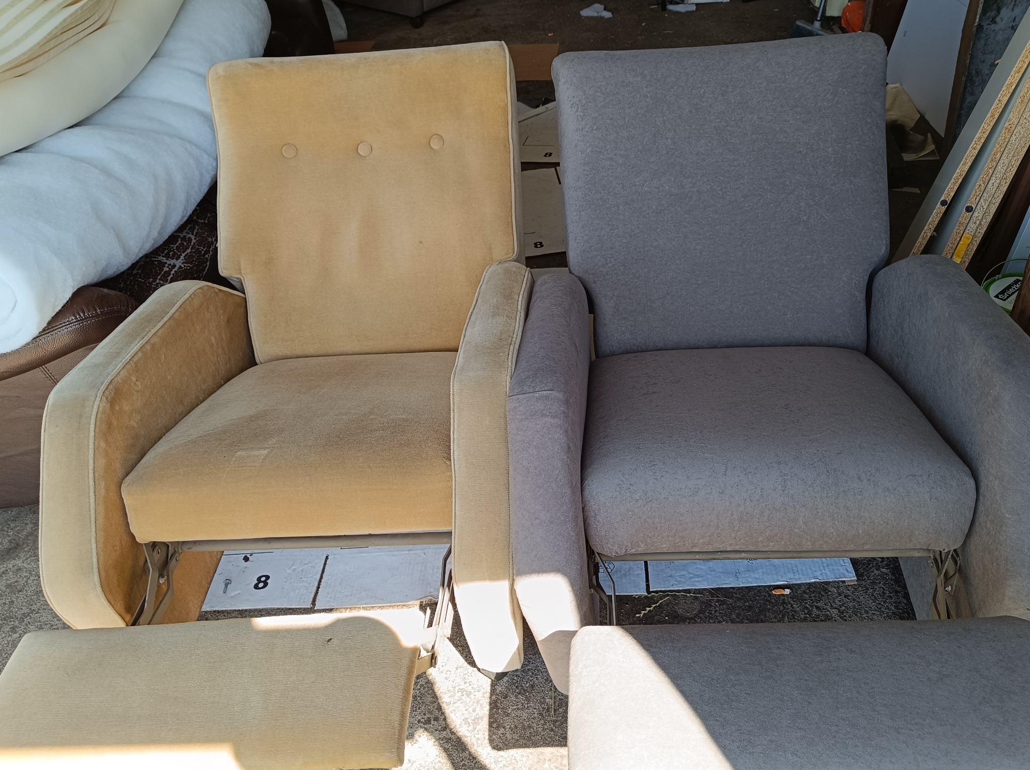 Dwa fotele obok siebie, jeden z beżową tapicerką z dwoma guzikami na oparciu, drugi z szarą tapicerką, oba z podnóżkami, widoczne elementy konstrukcyjne i kartonowe podkładki z numerem '8'.