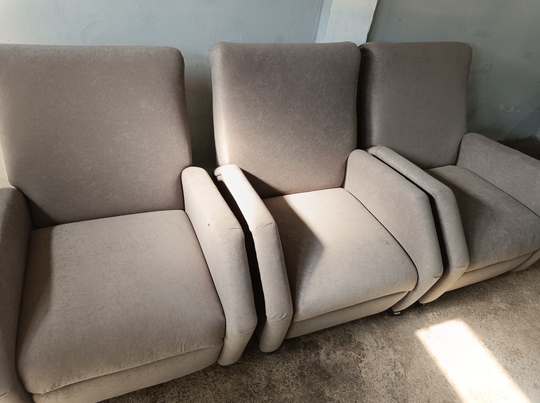 Trzy beżowe, tapicerowane fotele z podłokietnikami ustawione obok siebie, widoczne na tle szarej ściany i betonowej podłogi.