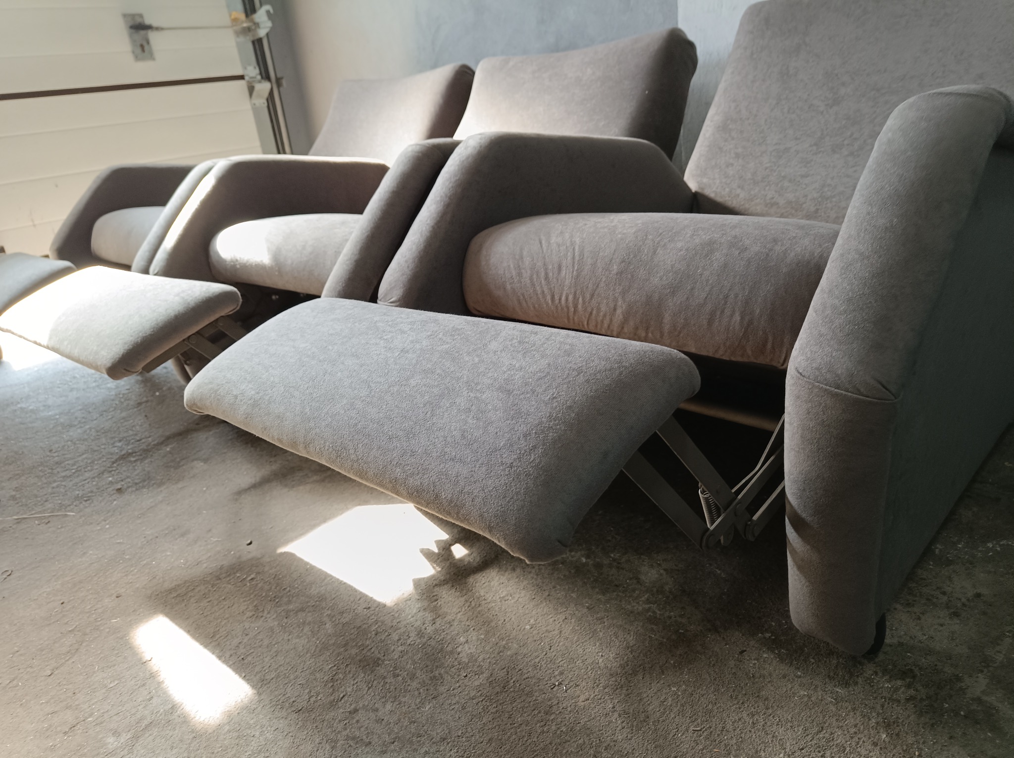 Rząd szarych, tapicerowanych foteli z wysuwanymi podnóżkami, ustawionych na betonowej podłodze, z widocznym mechanizmem rozkładania podnóżka.