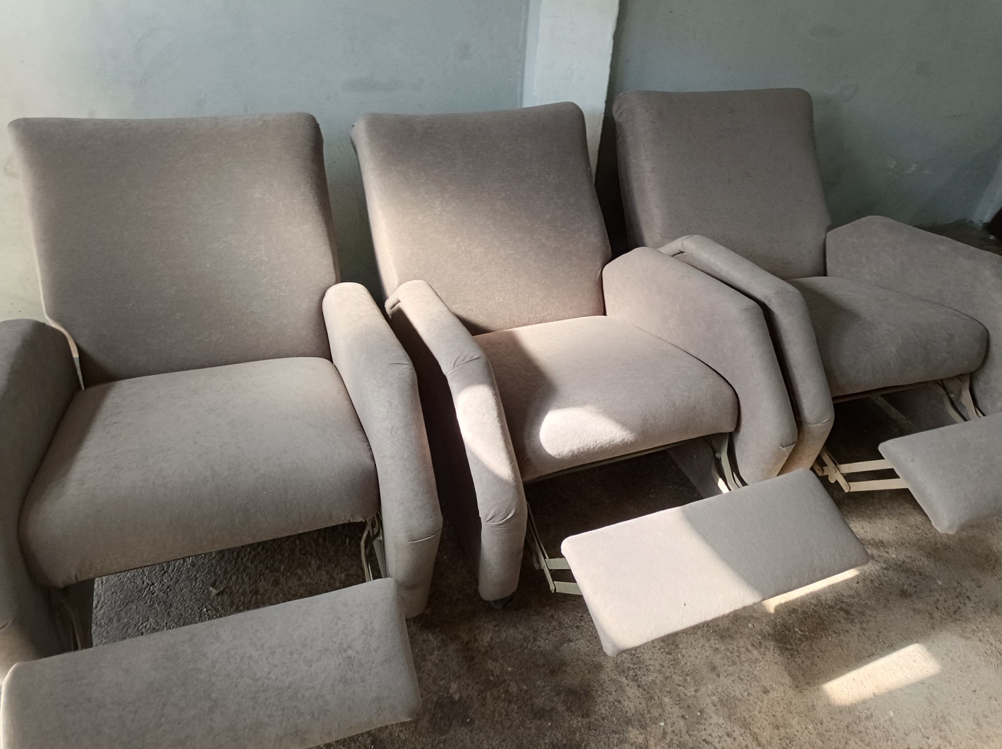 Trzy identyczne, beżowe fotele z podnóżkami, ustawione obok siebie w surowym wnętrzu, oświetlone naturalnym światłem padającym z góry.