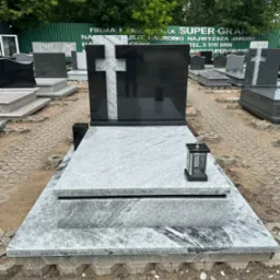 Granitowy pomnik z czarnym krzyżem i lampionem, widoczny na ekspozycji wśród innych nagrobków na zewnątrz.