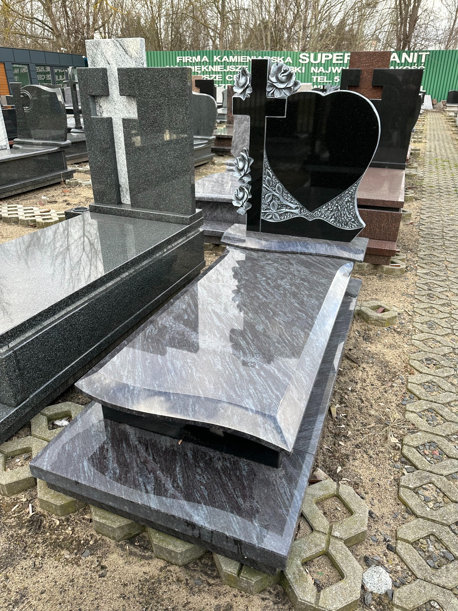 Wystawa nagrobków z ciemnego granitu, jeden z krzyżem i ornamentami w kształcie róż, drugi z prostym krzyżem, ustawione na brukowanym placu.
