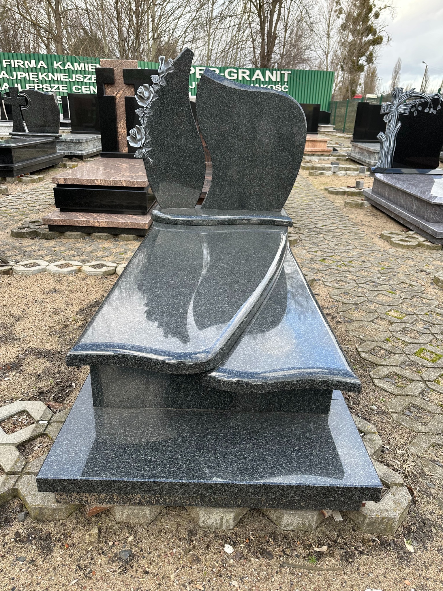 Czarny, granitowy nagrobek z motywem kwiatowym w kształcie łzy, prezentowany na placu kamieniarskim w pochmurny dzień.