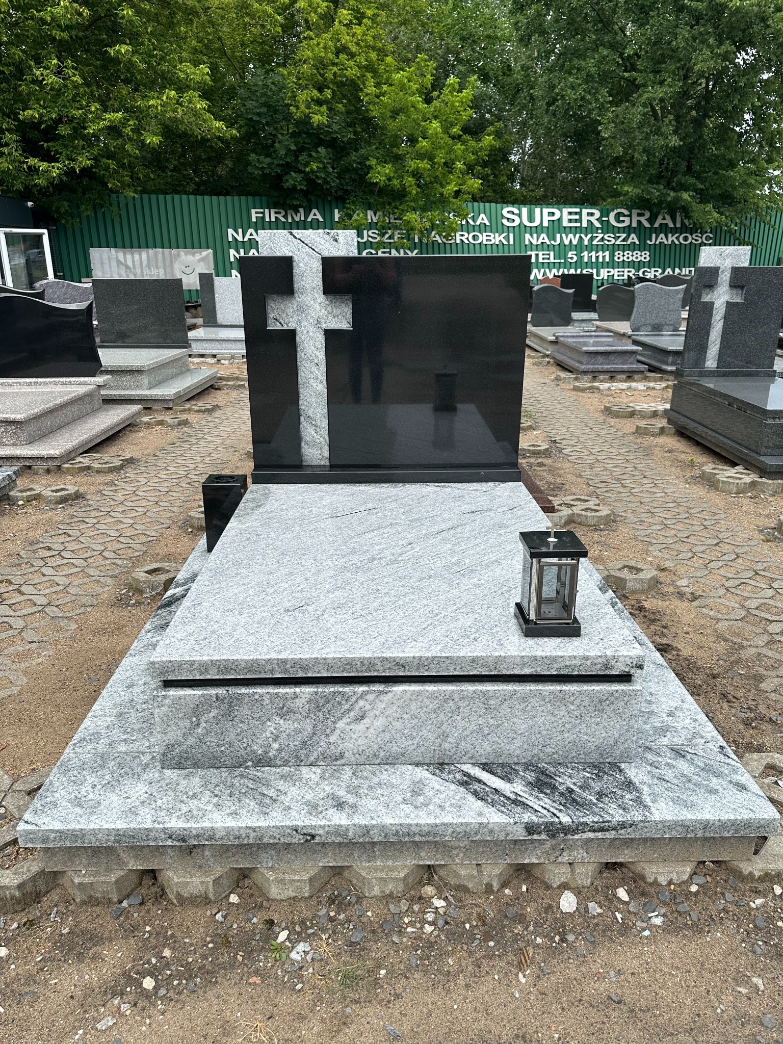 Granitowy pomnik z czarnym krzyżem i lampionem, widoczny na ekspozycji wśród innych nagrobków na zewnątrz.