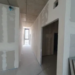 Korytarz w trakcie prac wykończeniowych, ściany pokryte szpachlą, widoczne otwory drzwiowe i okno w oddali, surowy betonowy sufit z wystającą instalacją.