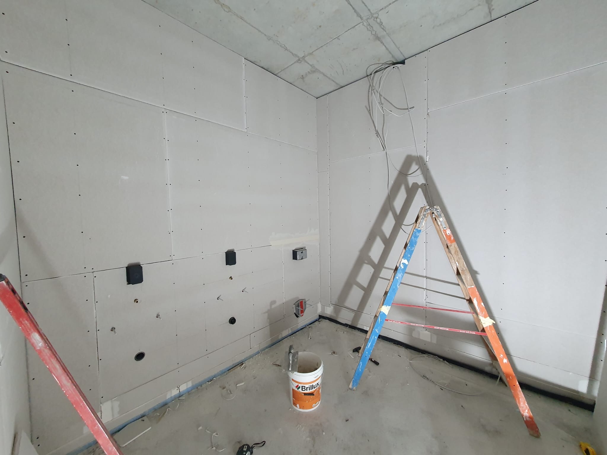 Wnętrze w trakcie montażu płyt kartonowo-gipsowych na ścianach i suficie, widoczne kable, puszki elektryczne, drabina oparta o ścianę oraz wiadro z farbą marki Brillux na podłodze.