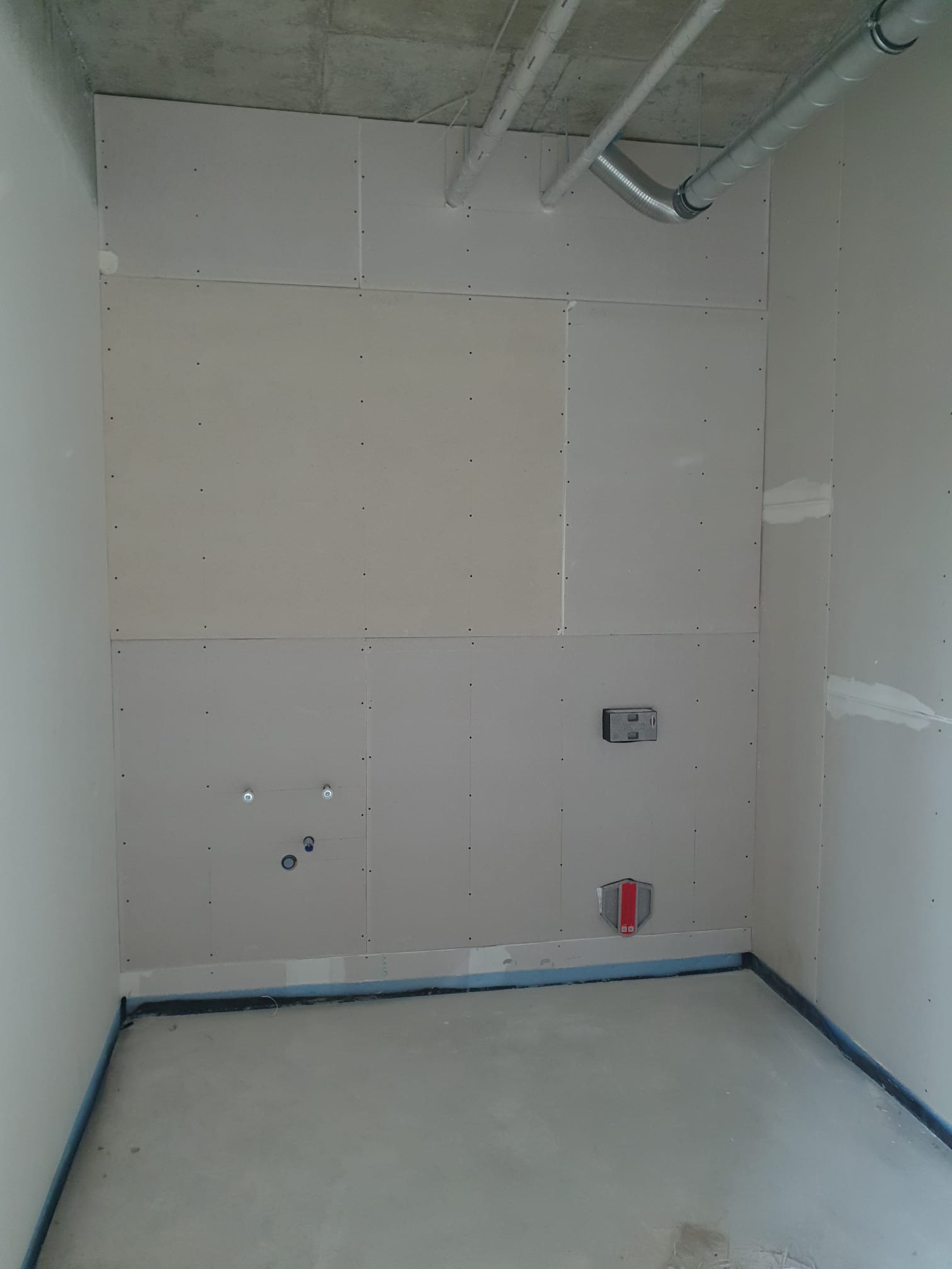 Surowe wnętrze w trakcie prac wykończeniowych: ściany obłożone płytami kartonowo-gipsowymi, widoczne instalacje i rury pod sufitem, zabezpieczona podłoga, puszki elektryczne w ścianie.