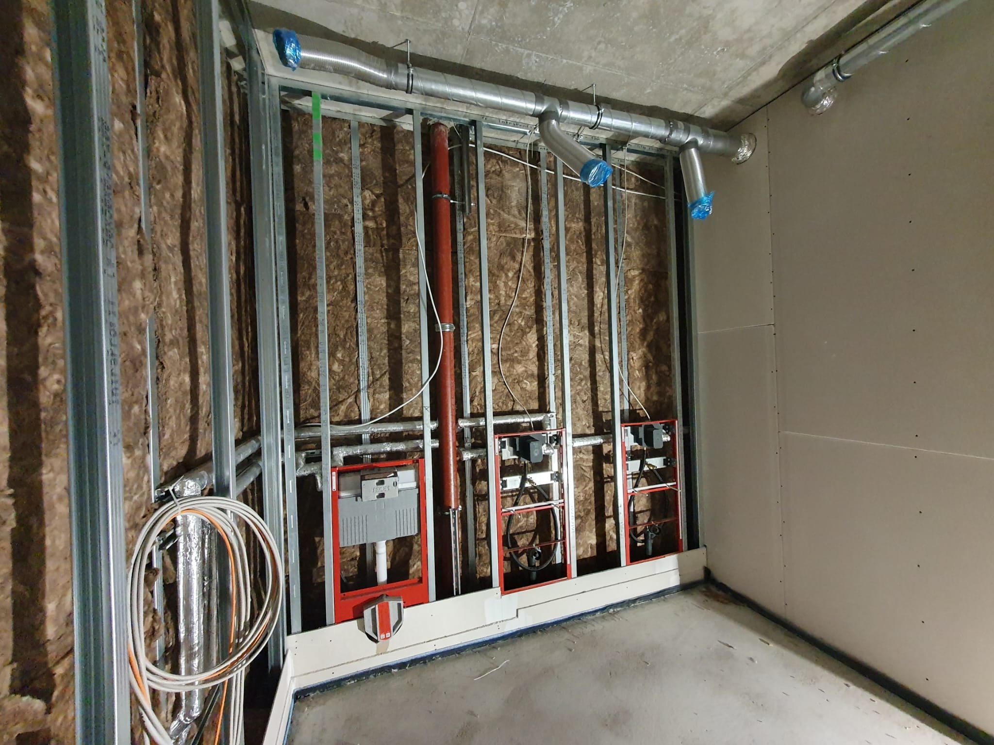 Wnętrze w trakcie prac wykończeniowych: widoczne metalowe profile konstrukcyjne, izolacja z wełny mineralnej, rury wentylacyjne, instalacje sanitarne w czerwonych ramach oraz fragment ściany...