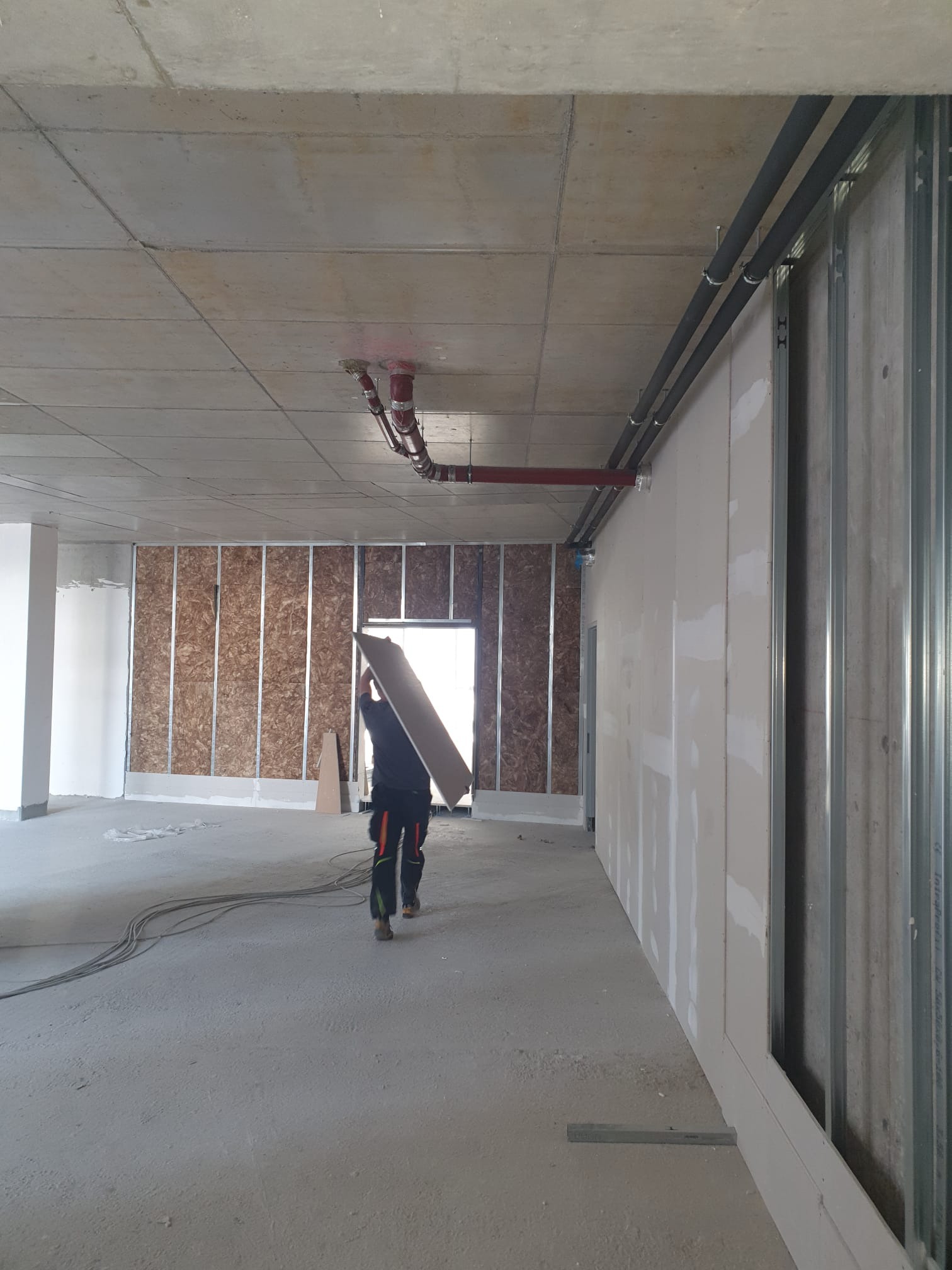 Pracownik w kombinezonie transportuje płytę gipsowo-kartonową w surowym wnętrzu, widoczne metalowe profile ścian działowych, instalacje pod sufitem i fragment ściany z płyt OSB.