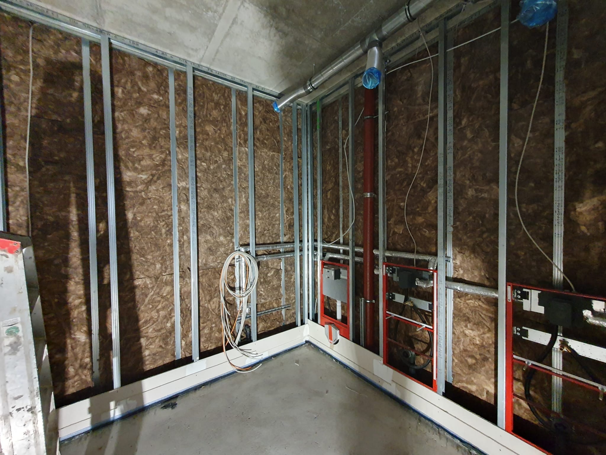 Wnętrze w trakcie prac: metalowy szkielet konstrukcji ścian działowych wypełniony brązową wełną mineralną, widoczne instalacje sanitarne i elektryczne, surowy betonowy sufit i podłoga.