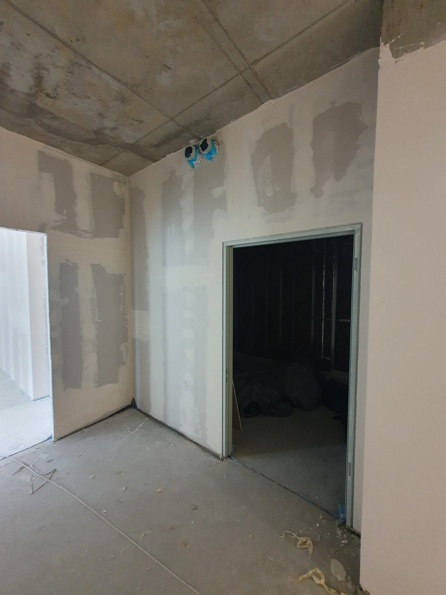 Surowy stan wnętrza w trakcie remontu: ściany pokryte warstwą szpachli, widoczne otwory na instalacje elektryczne w suficie, przygotowane do dalszych prac wykończeniowych.