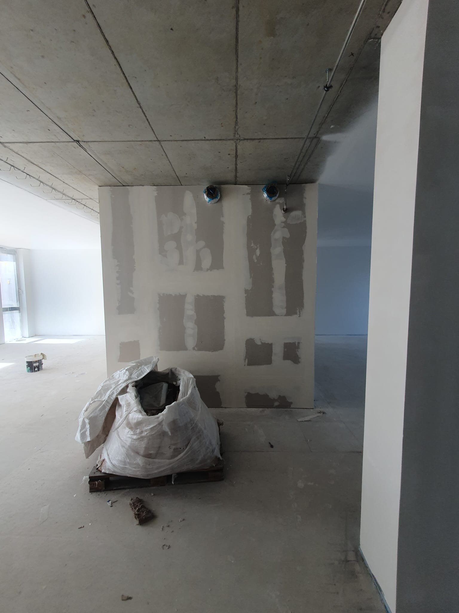 Wnętrze w trakcie prac w Miastku: ściana z płyt kartonowo-gipsowych częściowo pokryta szpachlą, worek z odpadami na palecie, surowy betonowy sufit z instalacjami.