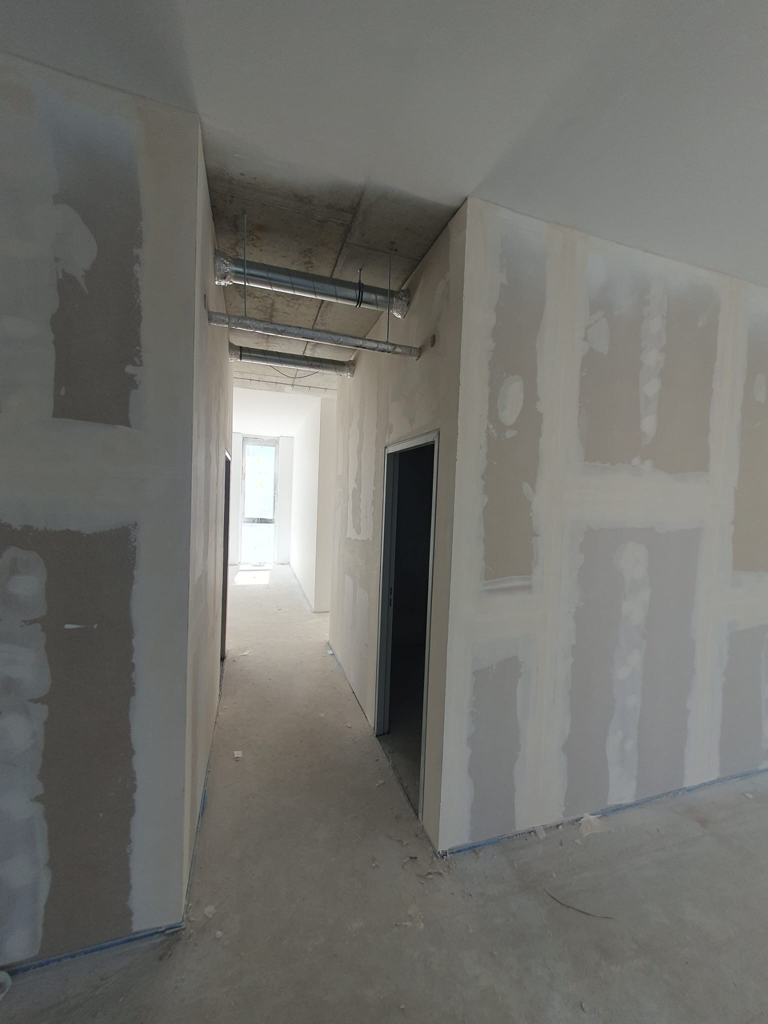 Korytarz w trakcie prac wykończeniowych; ściany z płyt kartonowo-gipsowych częściowo pokryte szpachlą, widoczne instalacje pod sufitem.