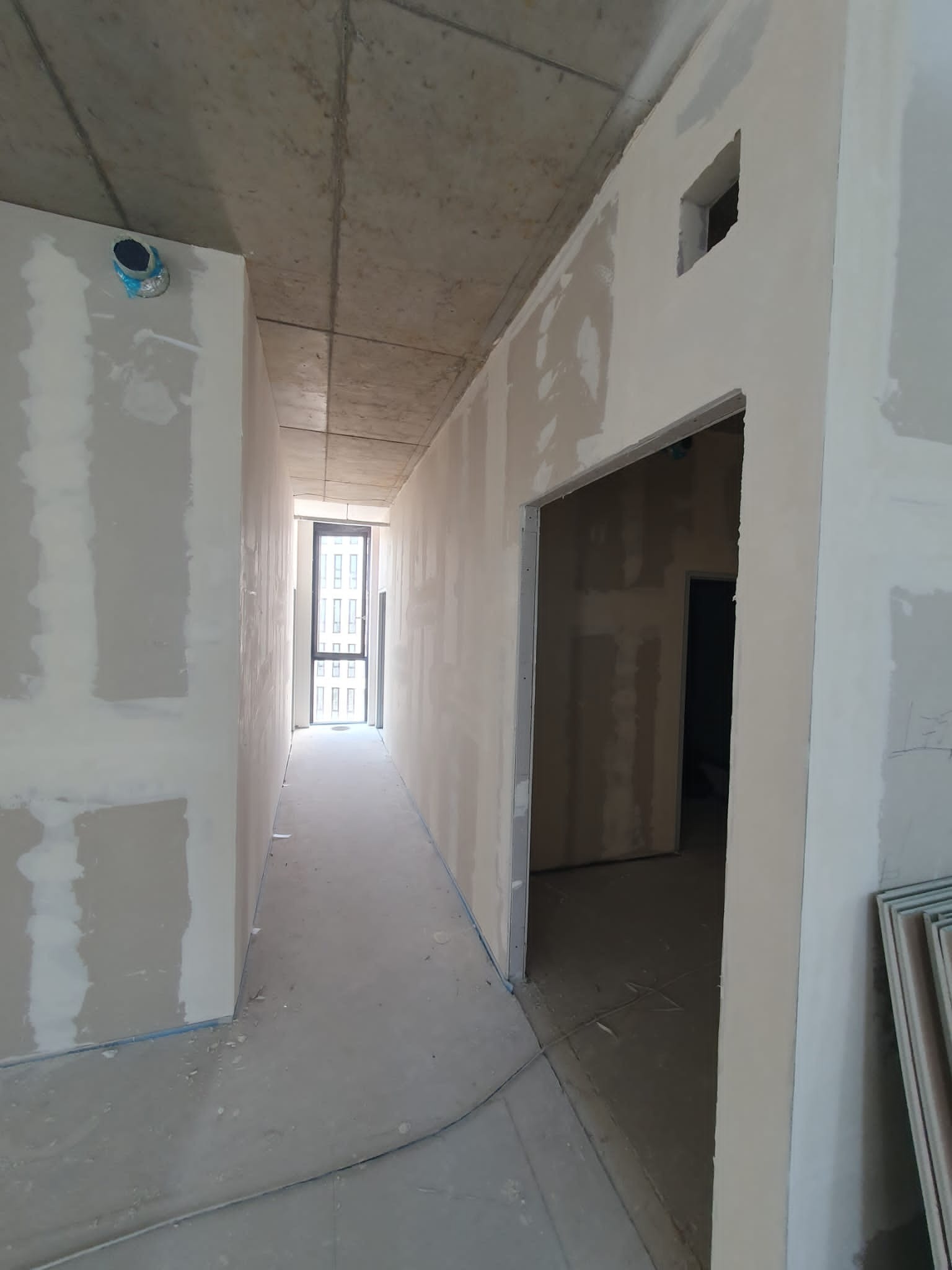 Korytarz w trakcie prac wykończeniowych, ściany pokryte szpachlą, widoczne otwory drzwiowe i okno w oddali, surowy betonowy sufit z wystającą instalacją.
