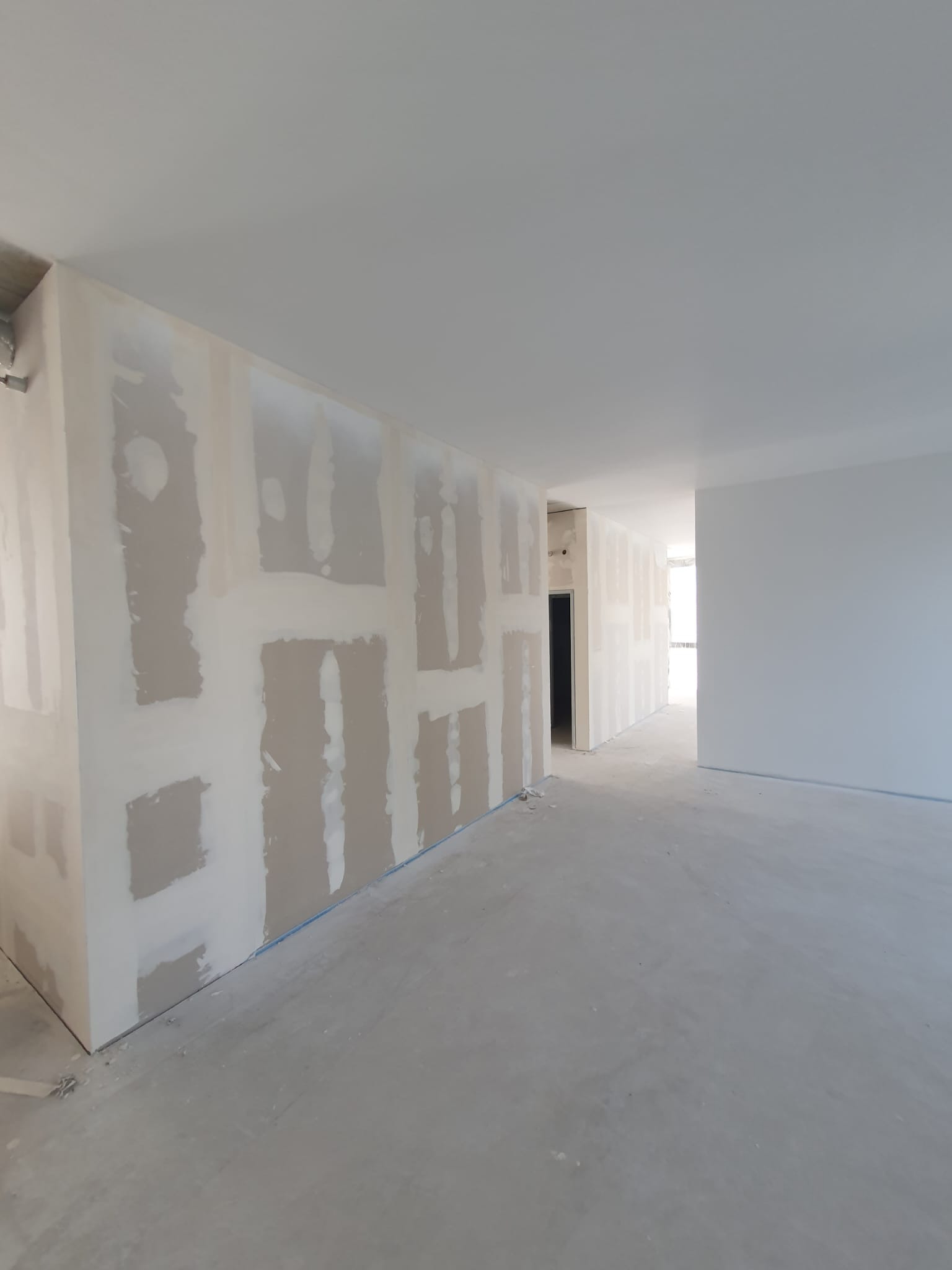 Surowe wnętrze z płytami gipsowo-kartonowymi na ścianach, częściowo pokrytymi masą szpachlową, surowa podłoga, widok na korytarz.