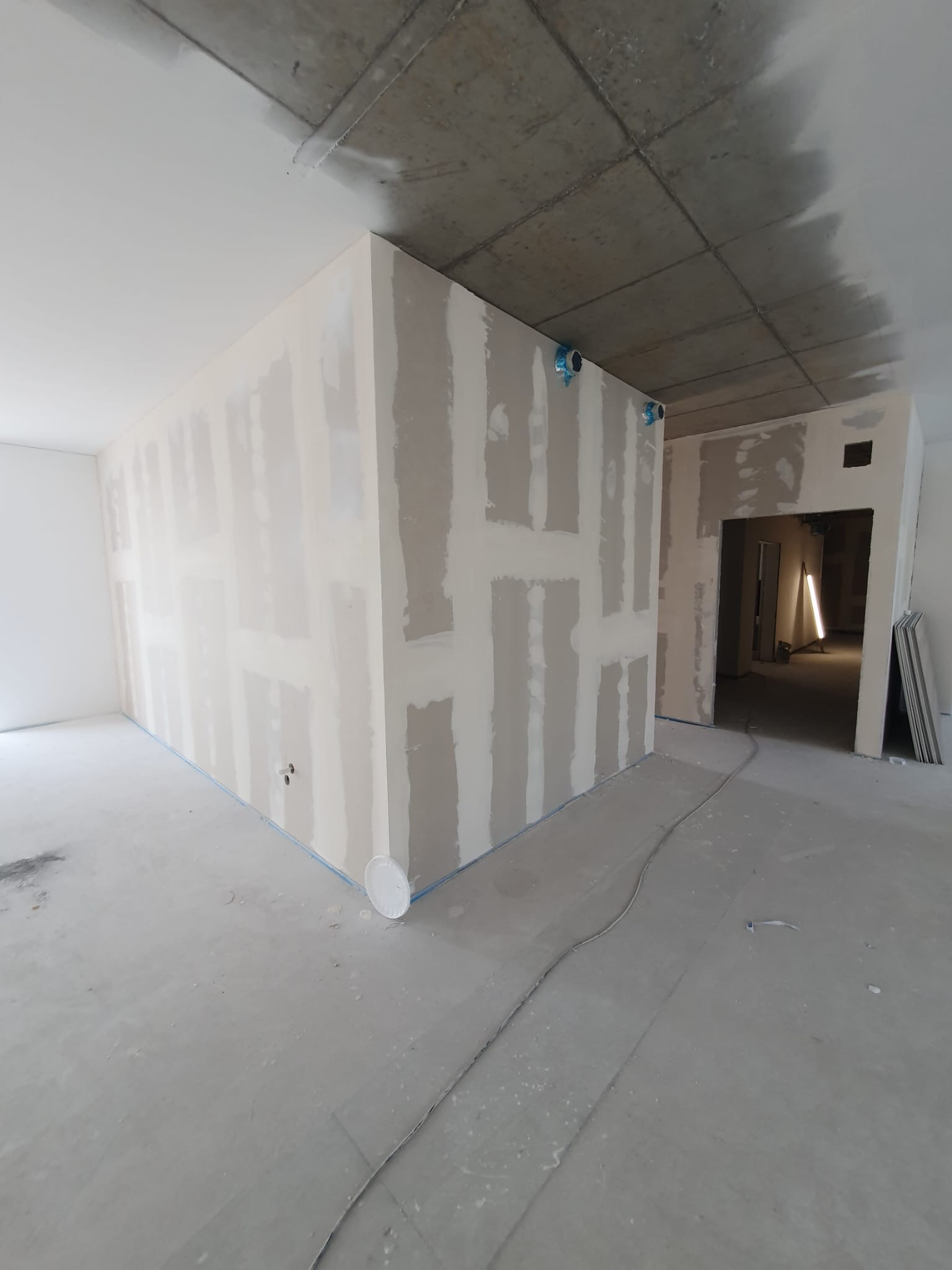 Wnętrze w trakcie prac wykończeniowych: ściany z płyt kartonowo-gipsowych z widocznymi łączeniami, surowy betonowy sufit, wystające rury instalacyjne zabezpieczone folią, szary betonowy podkład...