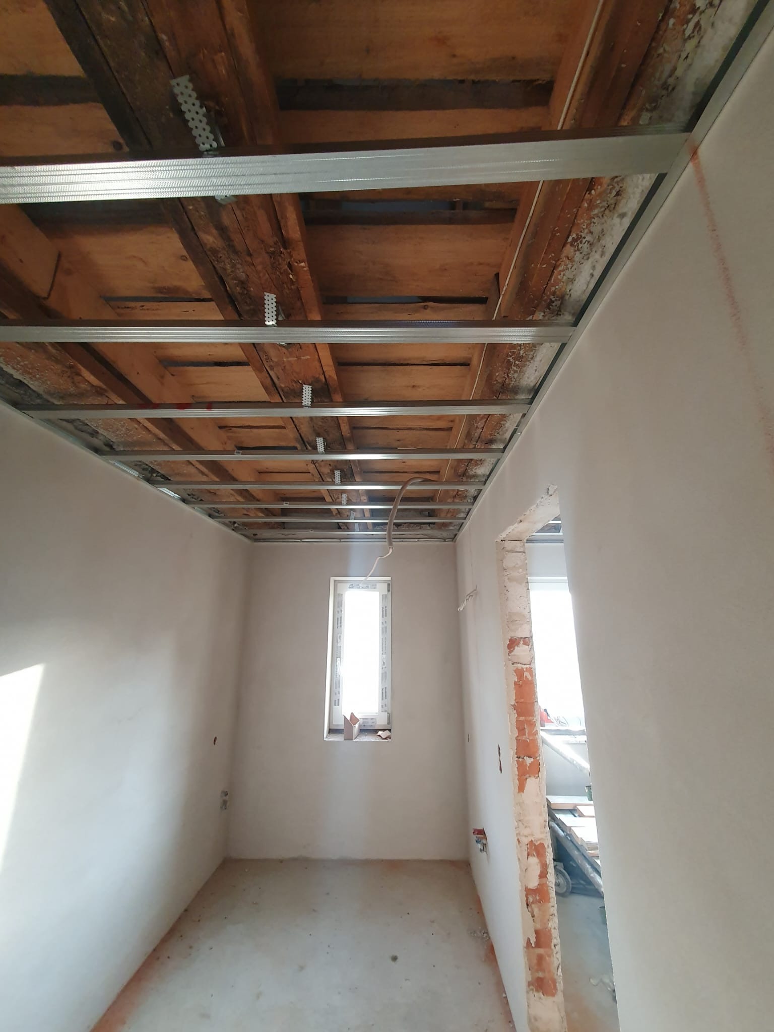 Wnętrze w trakcie montażu metalowej konstrukcji sufitu podwieszanego z widocznymi starymi drewnianymi belkami stropowymi, ściany po nałożeniu gładzi, okno w tle.