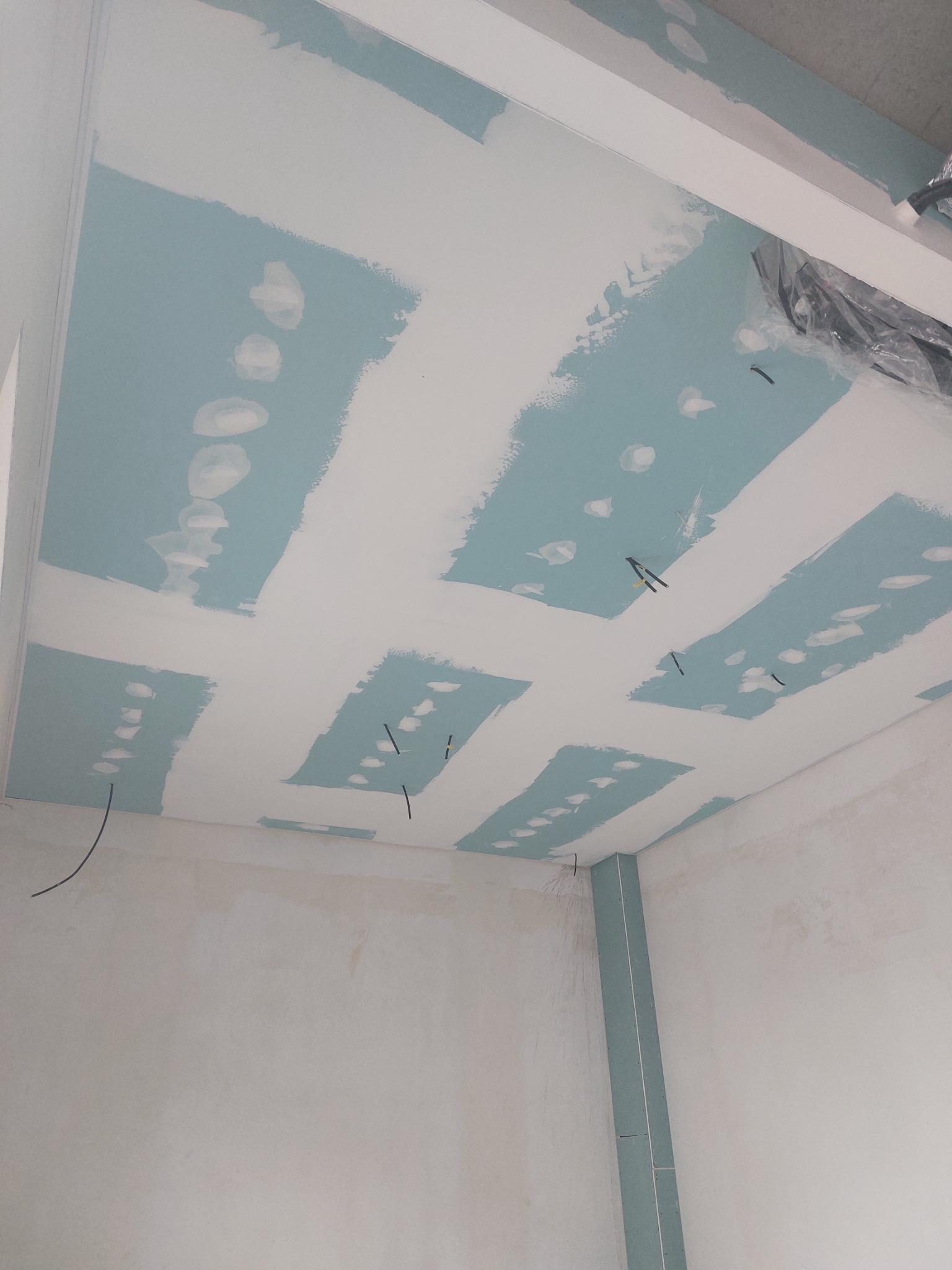 Niedokończony sufit z płyt kartonowo-gipsowych, widoczne łączenia płyt wypełnione białą masą szpachlową, surowe ściany w tle i wystające kable.