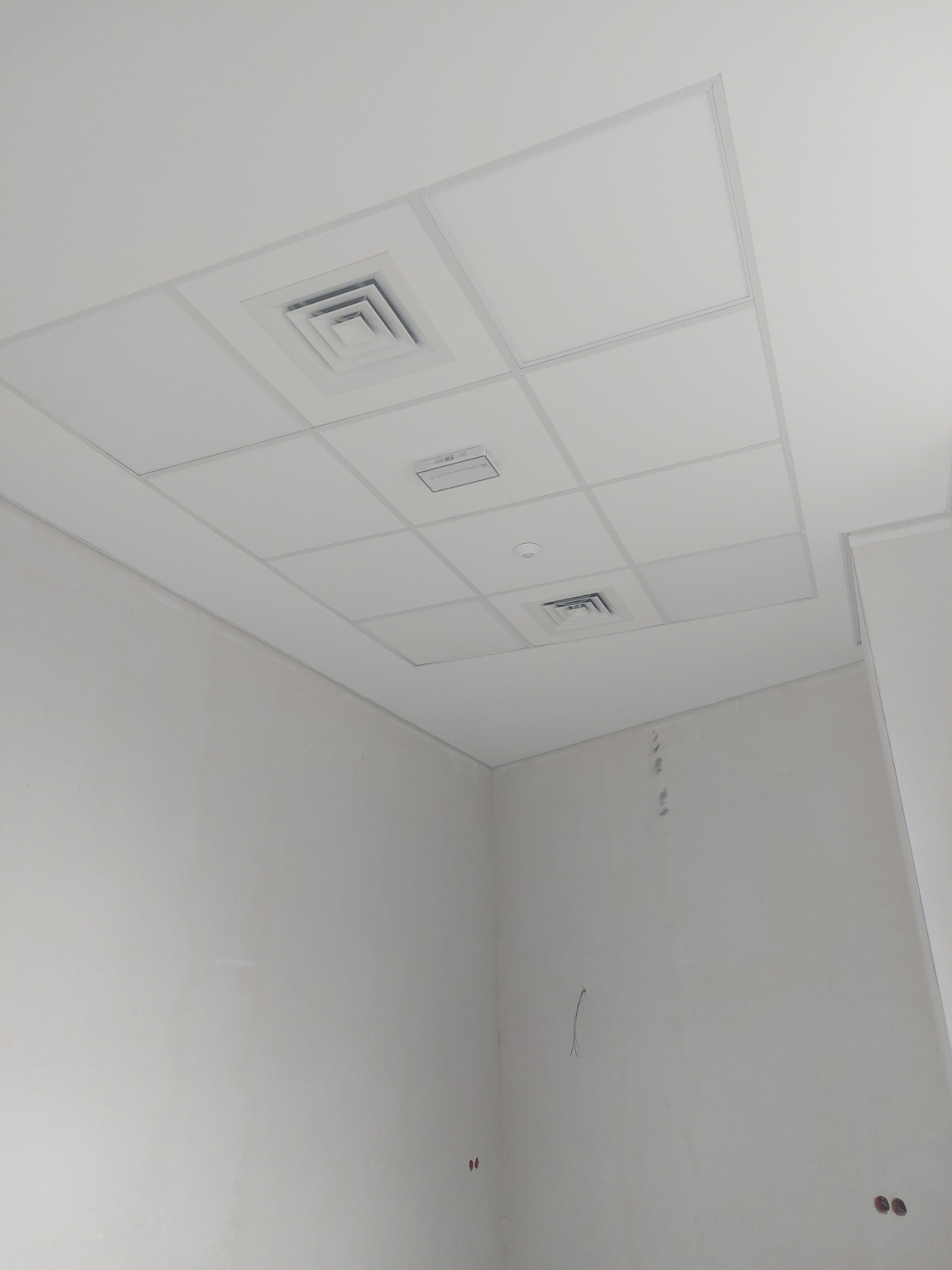 Kwadratowa, biała, podwieszana kaseta sufitowa z kratkami wentylacyjnymi w surowym, jasnym pomieszczeniu. Ściany w trakcie przygotowania, widoczne ślady po szpachlowaniu i wystające przewody.