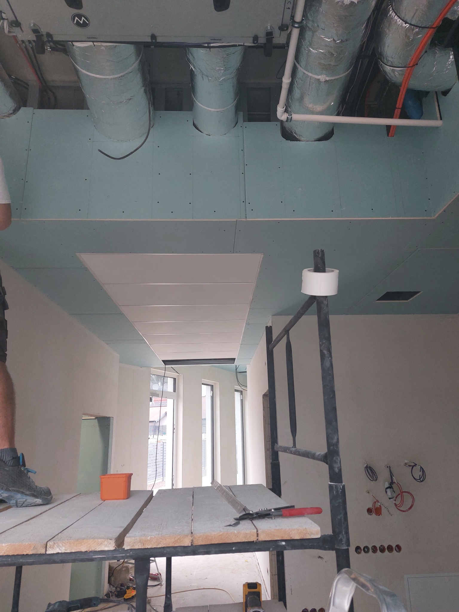 Montaż sufitu podwieszanego z płyt kartonowo-gipsowych, widoczne rury wentylacyjne pod stropem, rusztowanie z narzędziami i materiałami, instalacja elektryczna w ścianie, w tle okna.