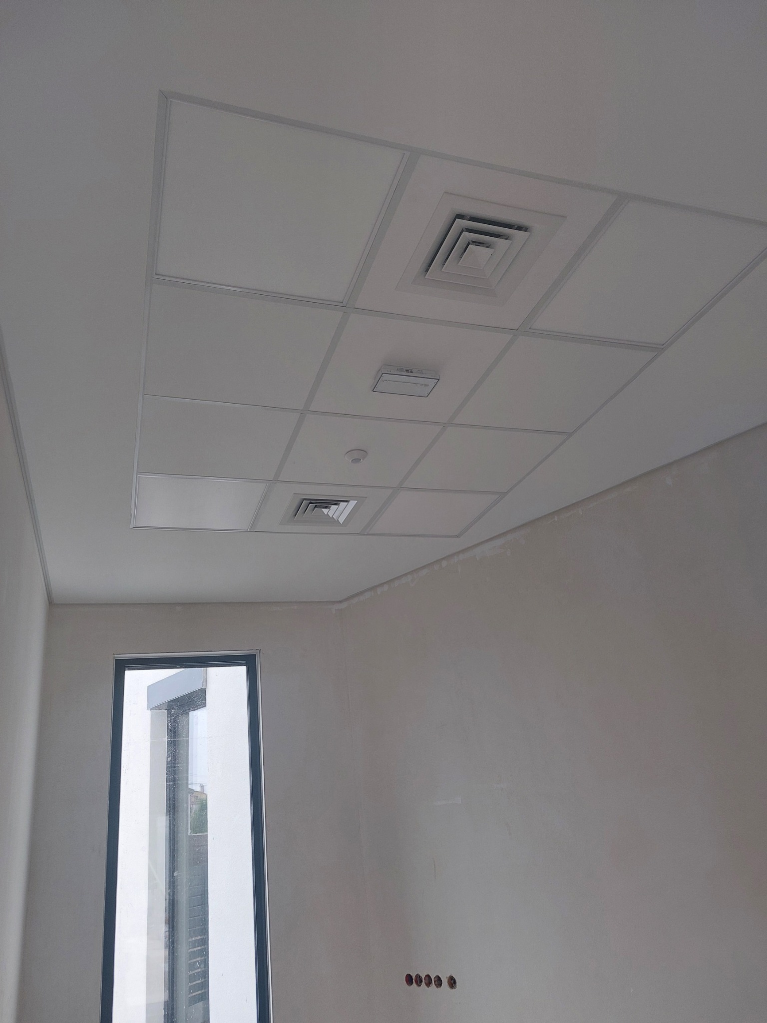 Sufit podwieszany z kwadratowymi panelami, kratkami wentylacyjnymi i oświetleniem w surowym wnętrzu z oknem i niezakończonymi ścianami.