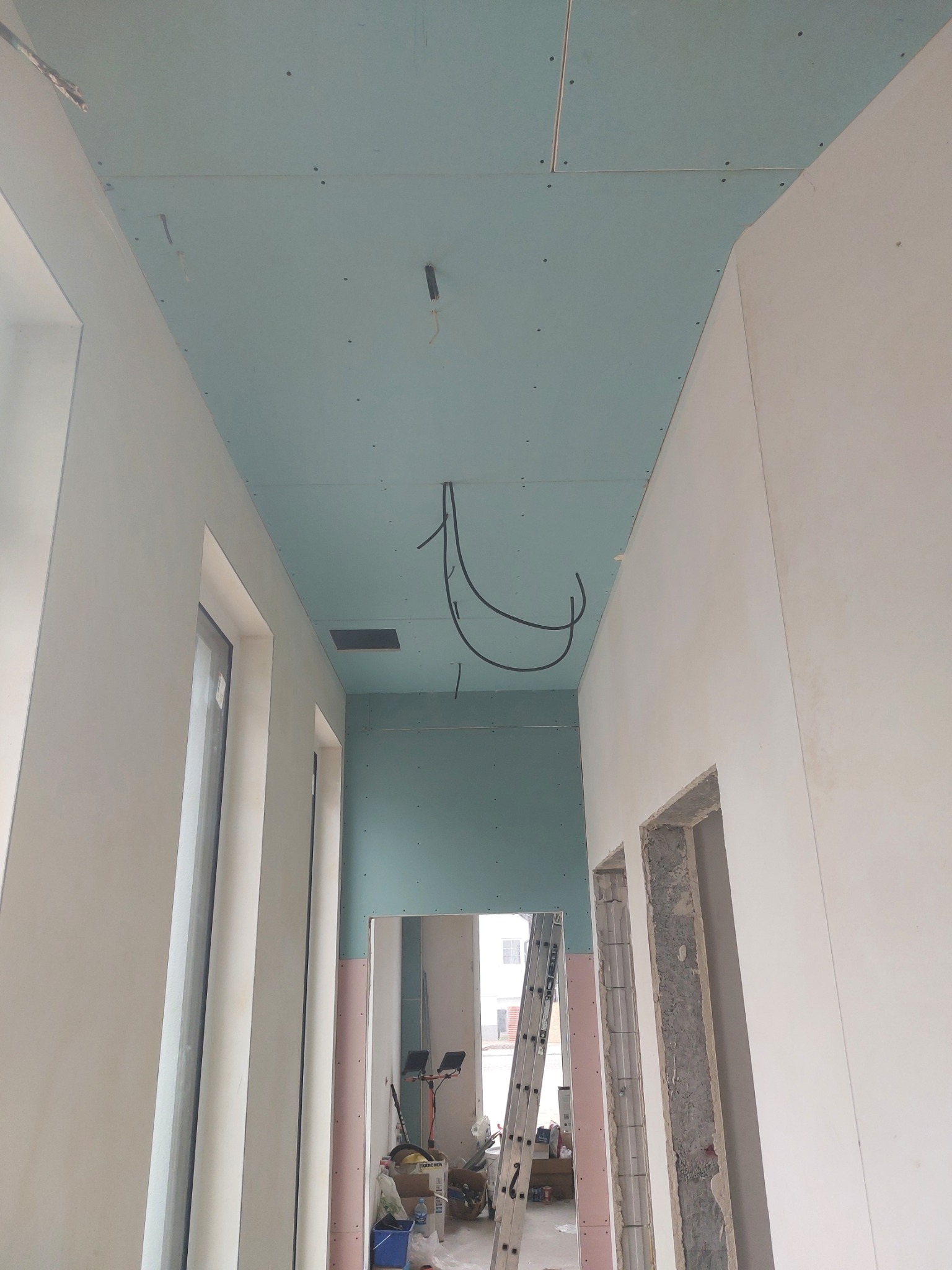 Korytarz w trakcie prac wykończeniowych: zamontowana płyta kartonowo-gipsowa na suficie, widoczne kable, ściany przygotowane pod malowanie, otwory drzwiowe w surowym stanie, drabina i narzędzia w tle.
