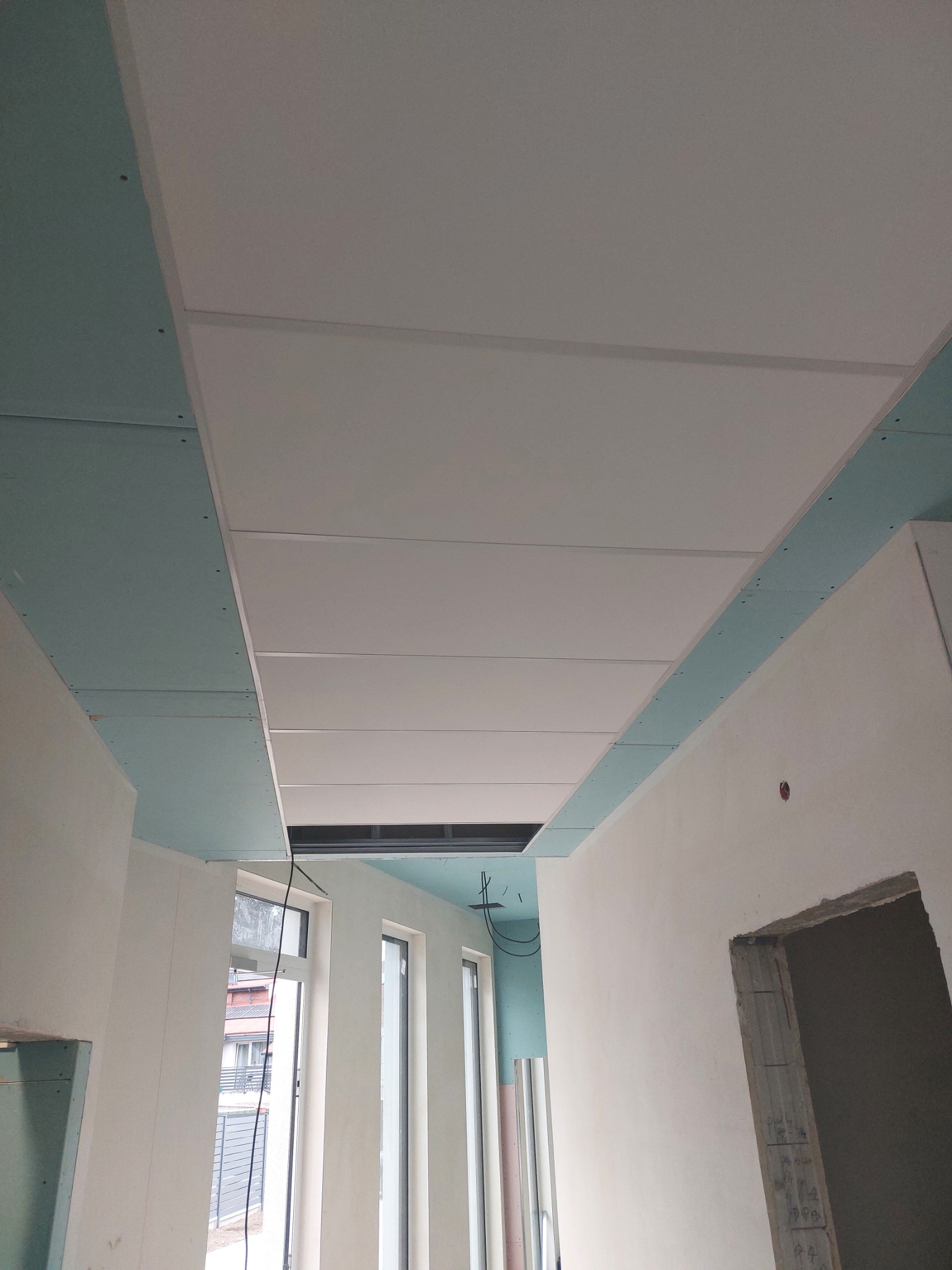 Montaż sufitu podwieszanego z płyt kartonowo-gipsowych, widoczne profile i płyty w trakcie instalacji, surowe ściany w tle, okna z widokiem na zewnątrz.