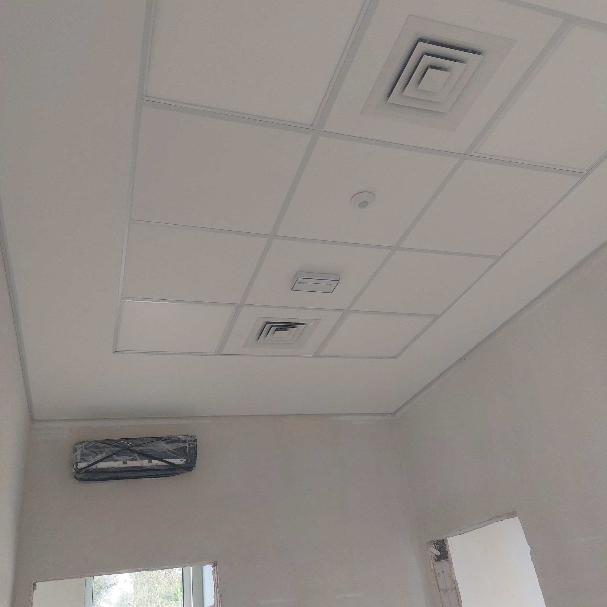 Kasetonowy sufit podwieszany z kratką wentylacyjną i czujnikiem dymu, klimatyzator zabezpieczony folią, ściany w trakcie prac wykończeniowych, widoczny fragment okna.