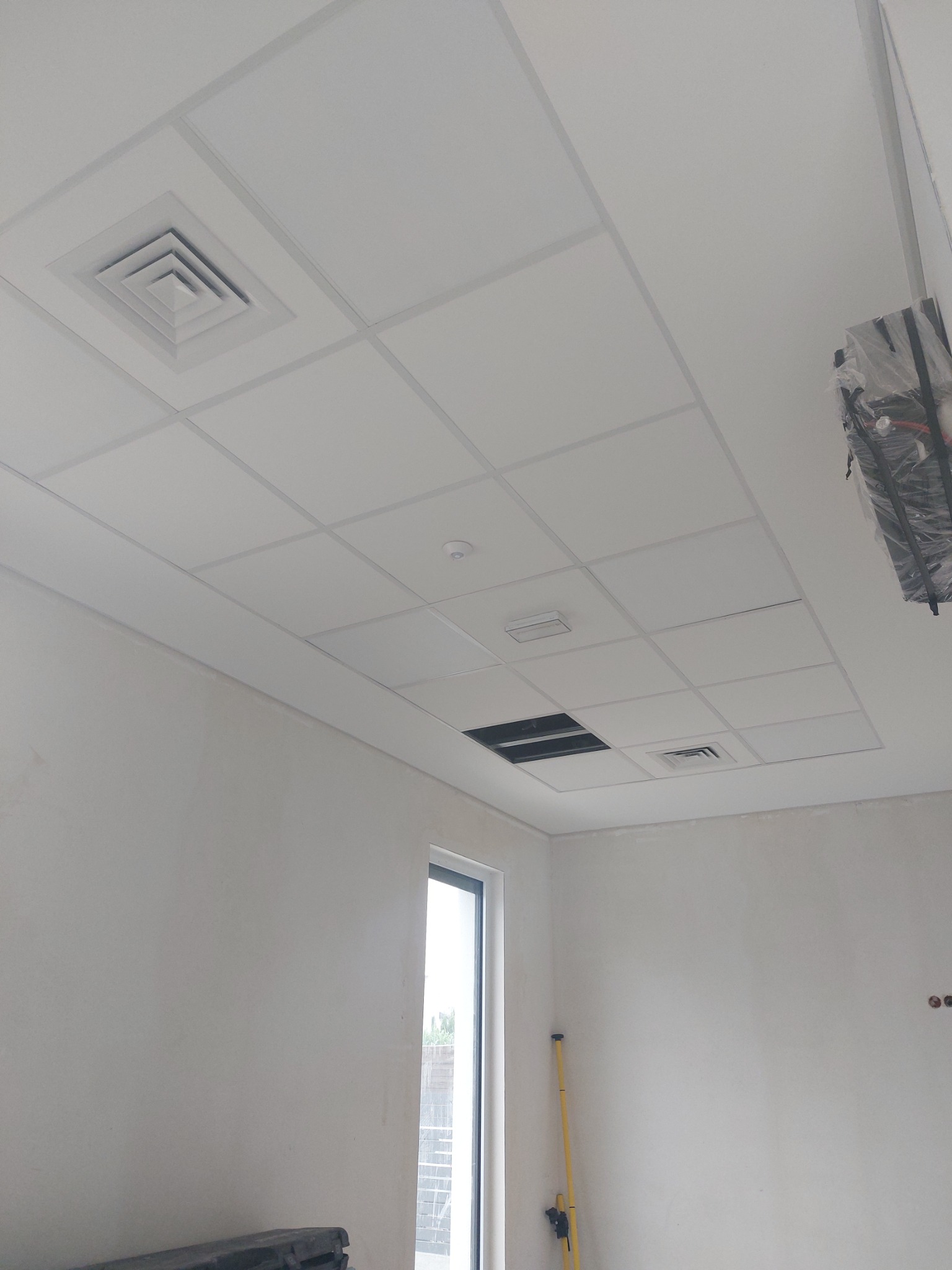 Wnętrze w trakcie prac wykończeniowych, widoczny sufit podwieszany kasetonowy z kratką wentylacyjną, ściany przygotowane do malowania, okno w tle, elementy instalacji zabezpieczone folią.
