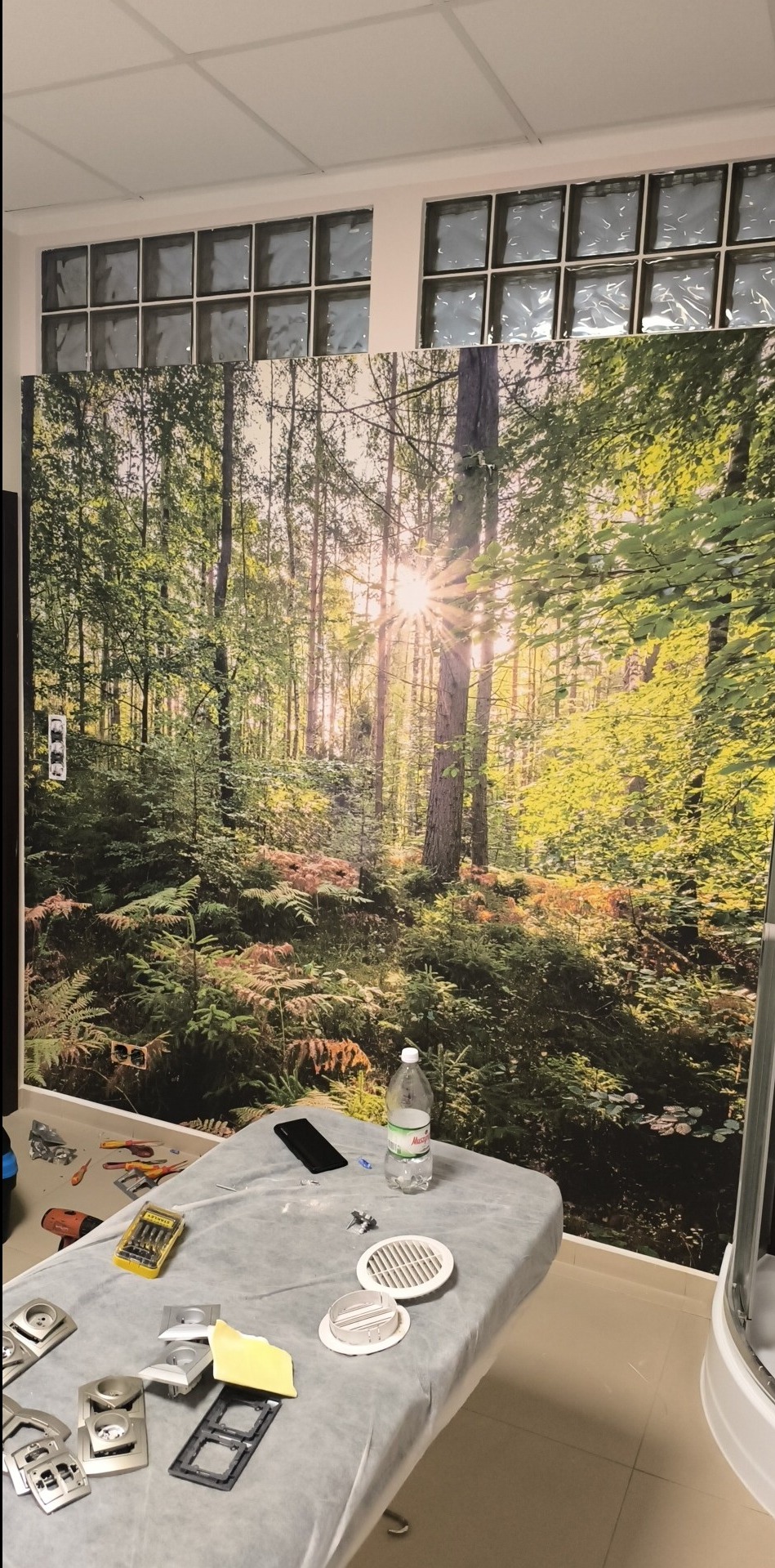 Fototapeta z motywem leśnym w trakcie montażu, widoczne narzędzia, elementy wykończeniowe, butelka wody i telefon na stole przy ścianie z fototapetą, okna z luksferami w górnej części ściany.