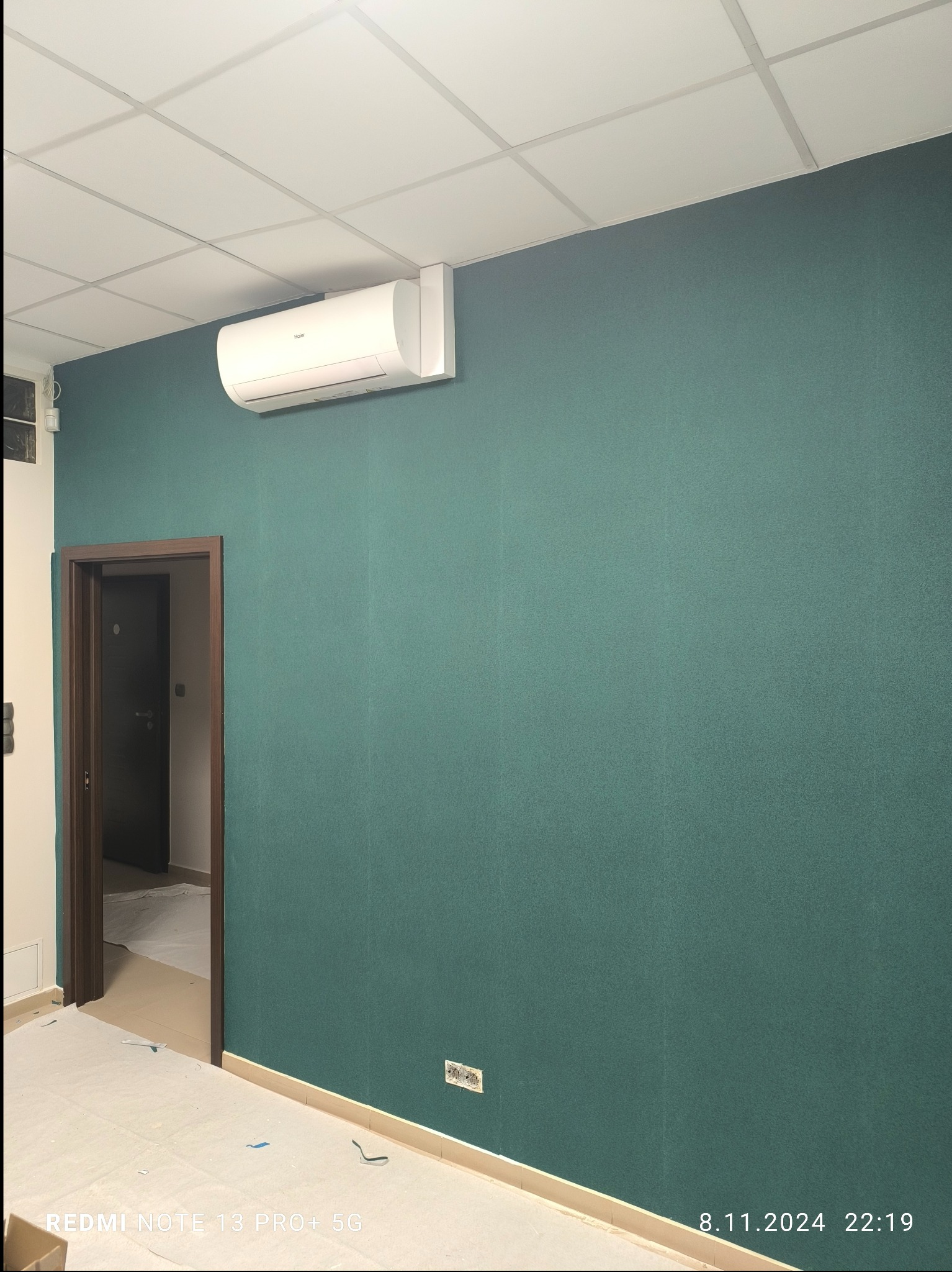 Ściana w odcieniu butelkowej zieleni z zamontowanym klimatyzatorem, widoczne gniazdko elektryczne i fragment podłogi zabezpieczonej folią malarską.