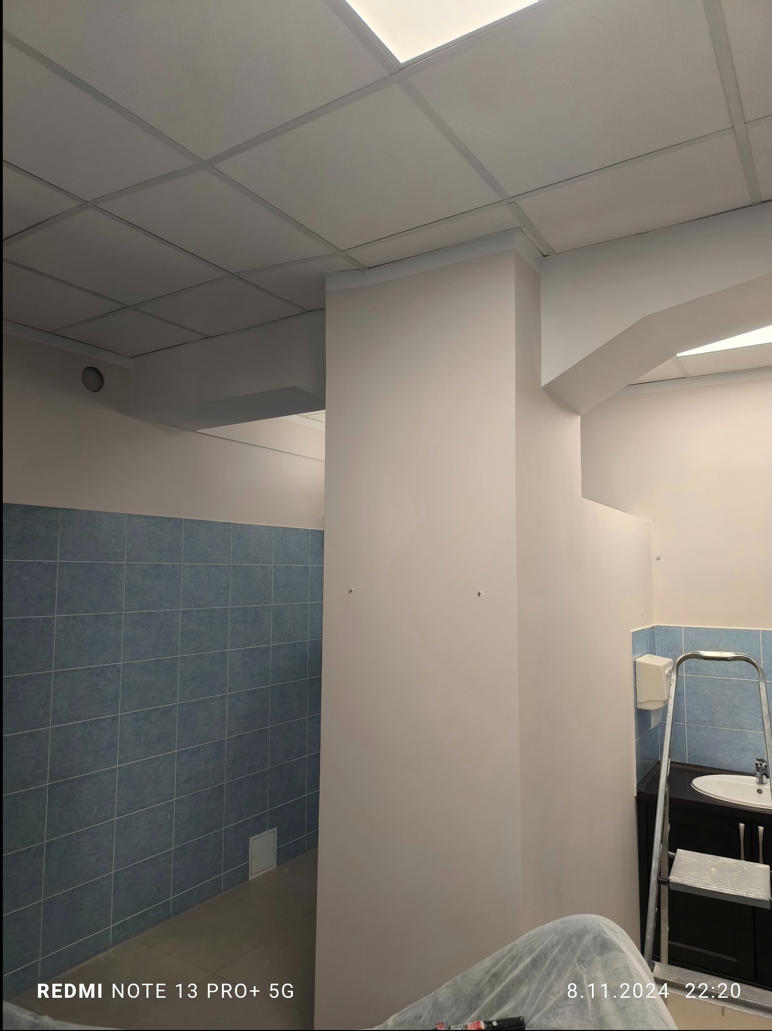 Świeżo pomalowane na biało ściany w pomieszczeniu z niebieskimi płytkami, widoczna drabina aluminiowa i fragment umywalki, sufit kasetonowy z oświetleniem.